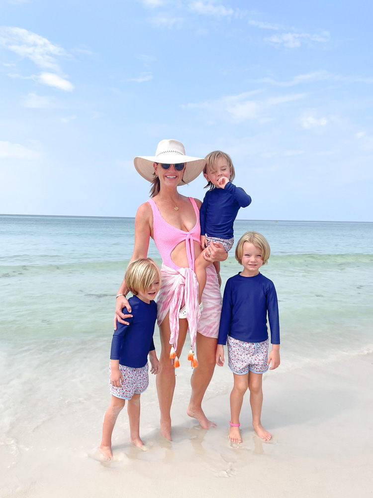 mom in pink swimsuit with three young sons in navy rash guards at the beach