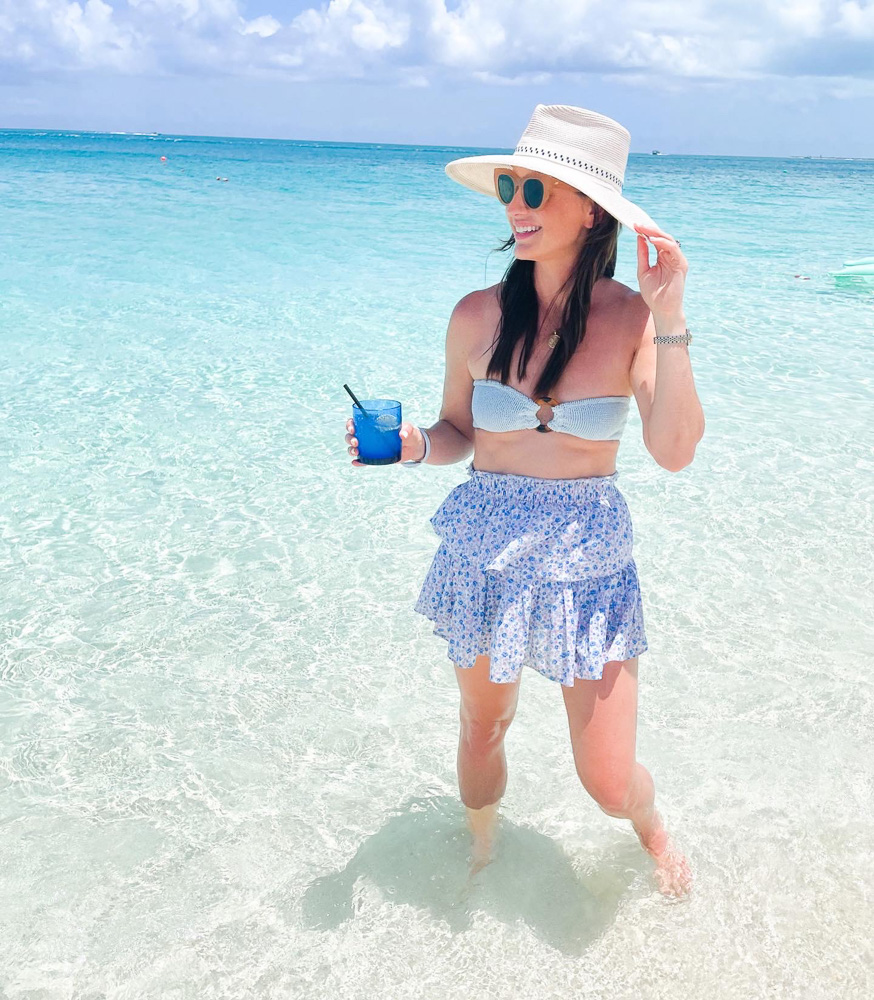 woman standing in swim suit and hat in ocean with a drink in hand