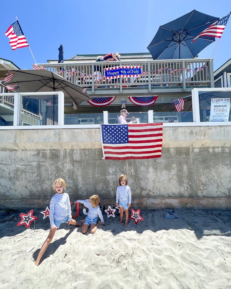 three little boys in swim suits standing under american flag at the beach