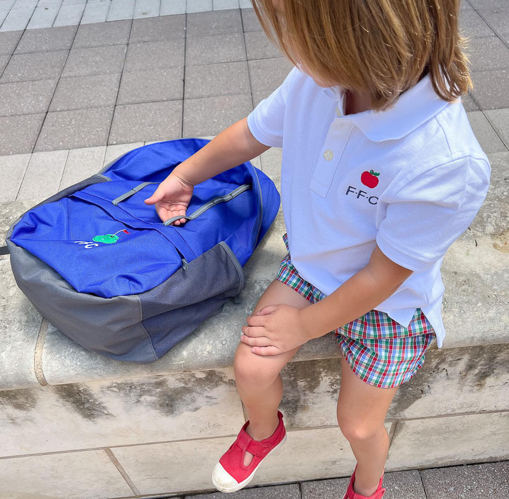 toddler sitting on bench with blue backpack wearing white polo with apple and initials monogram
