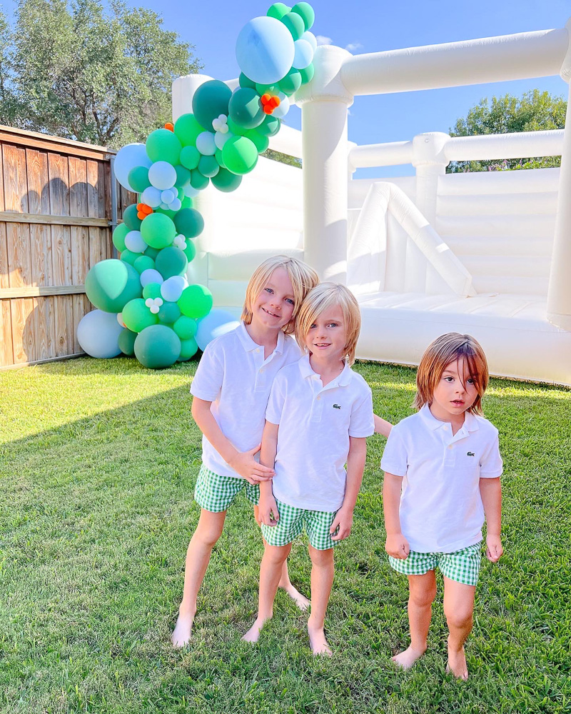 three little boys in matching shorts and polos standing in front of bounce house with green balloon garland
