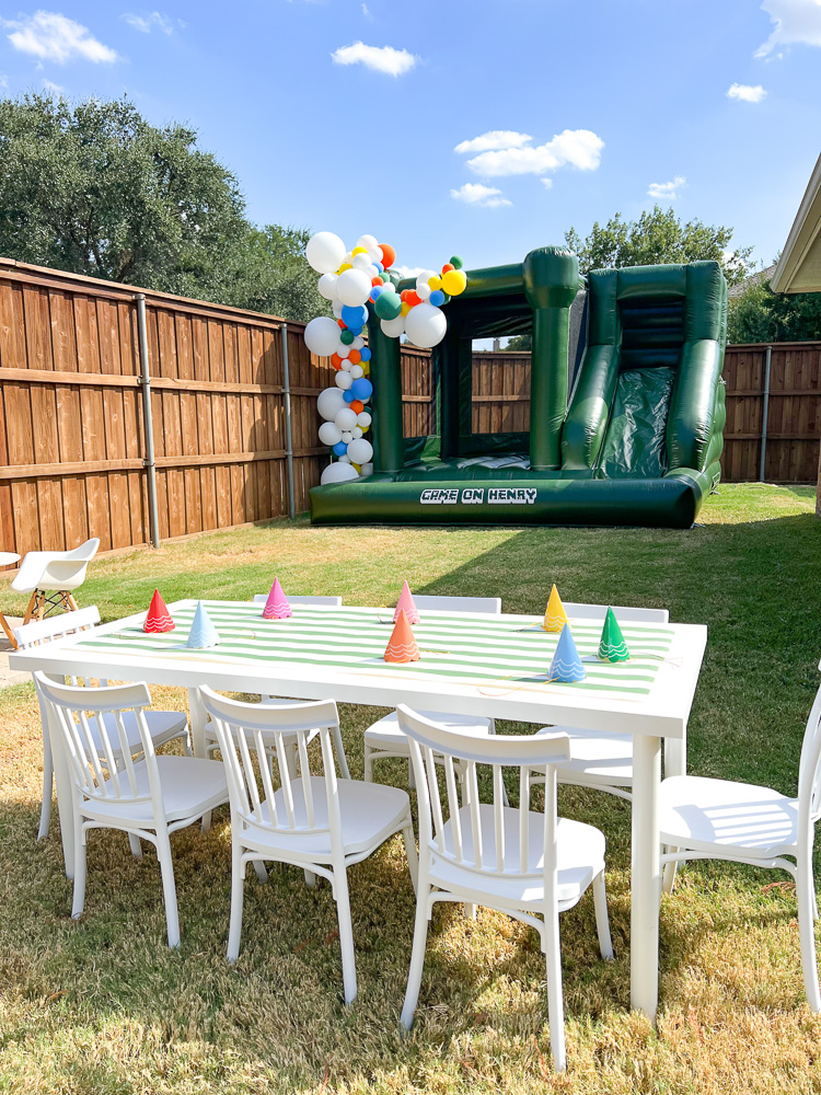 backyard setting for birthday party large green bounce house and white childrens table and chairs